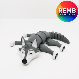 Puede incluir: Un juguete de lobo articulado de color gris y blanco con un cuerpo segmentado. El juguete está tumbado de lado con la cabeza apoyada en el suelo.