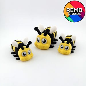 Può includere: Tre figurine di api dei cartoni animati gialle e nere con ali bianche e grandi occhi amichevoli. Le api sono disposte su uno sfondo bianco. Il logo REMB STUDIOS si trova nell'angolo in alto a destra.