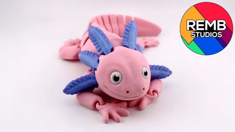 May include: A pink and blue axolotl figurine with articulated limbs and gills. The figurine has large, round eyes and is lying on a white surface. The REMB STUDIOS logo is in the background.