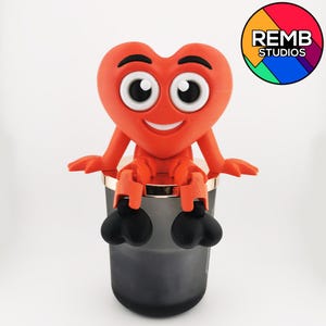 Puede incluir: Un personaje rojo en forma de coraz&oacute;n con una cara sonriente y ojos grandes se sienta en la parte superior de un tarro de vela negro. El personaje tiene brazos y piernas y se agarra al tarro con las manos. El tarro tiene un borde dorado.
