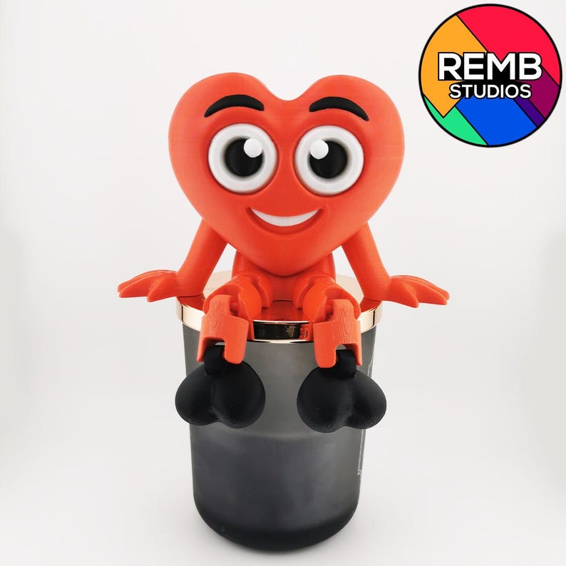 Puede incluir: Un personaje rojo en forma de coraz&oacute;n con una cara sonriente y ojos grandes se sienta en la parte superior de un tarro de vela negro. El personaje tiene brazos y piernas y se agarra al tarro con las manos. El tarro tiene un borde dorado.