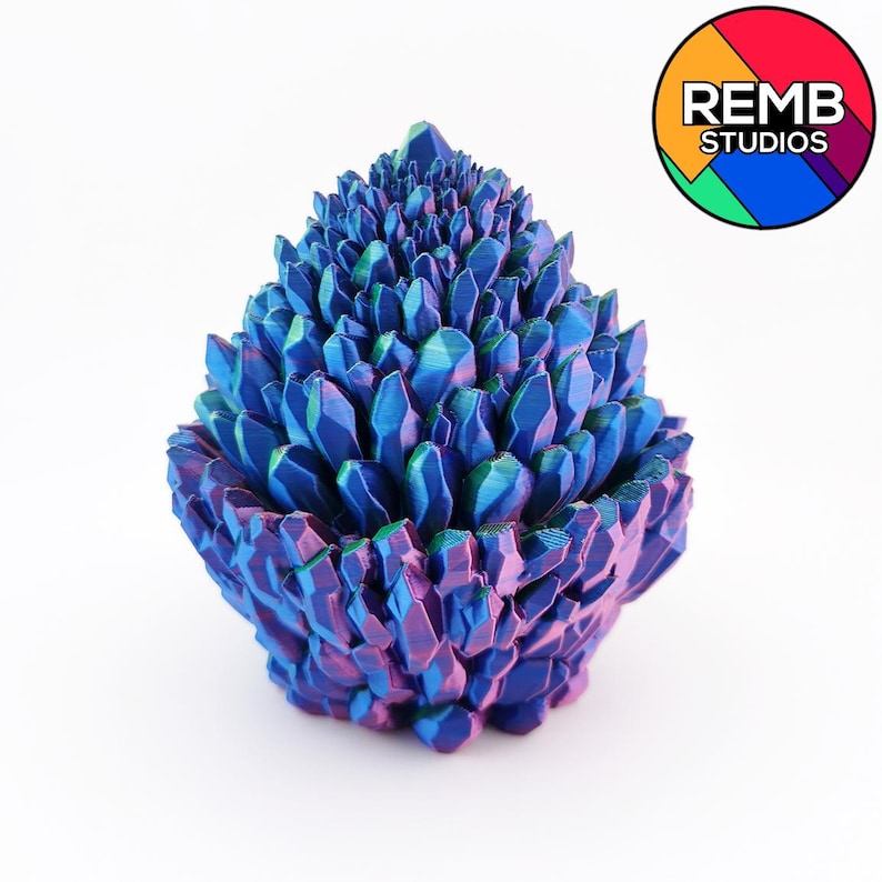 May include: A decorative, iridescent crystal cluster sculpture in shades of blue, purple, and green. The sculpture has a layered, textured appearance, with a rounded base and a pointed top. The REMB STUDIOS logo is in the upper right corner.