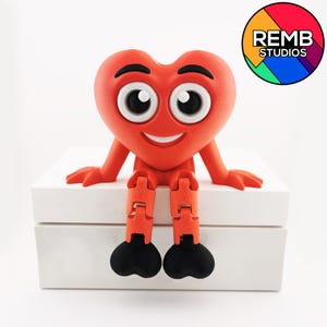 Puede incluir: Una figurita roja en forma de coraz&oacute;n con una cara sonriente y ojos grandes se sienta en una caja blanca. La figurita tiene piernas articuladas y pies negros. El texto "REMB STUDIOS" es visible en un c&iacute;rculo colorido en la esquina superior derecha de la imagen.