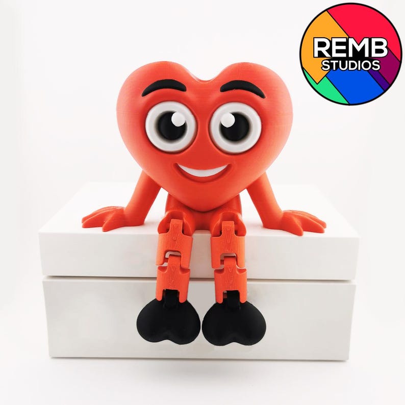 Puede incluir: Una figurita roja en forma de coraz&oacute;n con una cara sonriente y ojos grandes se sienta en una caja blanca. La figurita tiene piernas articuladas y pies negros. El texto "REMB STUDIOS" es visible en un c&iacute;rculo colorido en la esquina superior derecha de la imagen.