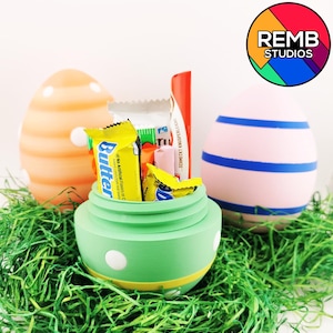 Puede incluir: Un huevo de Pascua verde con lunares blancos y una franja amarilla está abierto y lleno de caramelos. Los caramelos incluyen Butterfinger, una barra de caramelo rosa y blanca, y una barra de caramelo azul y amarilla. El huevo está sentado en un lecho de hierba verde. Hay otros dos huevos de Pascua en el fondo, uno naranja y otro rosa con rayas azules.