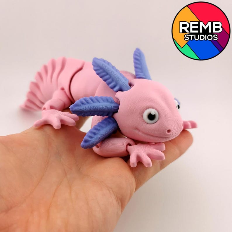 May include: A pink and blue axolotl figurine with detailed gills and limbs. The figurine is held in a hand, showcasing its articulated design. The axolotl has a detailed tail and a smiling expression. The logo "REMB STUDIOS" is in the background.