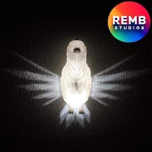 Könnte beinhalten: Eine weiße, keramikförmige Vogellampe mit strukturierter Oberfläche wirft einen Schatten von Flügeln an die Wand. Die Lampe ist beleuchtet und leuchtet.