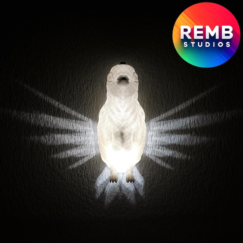 K&ouml;nnte beinhalten: Eine wei&szlig;e, keramikf&ouml;rmige Vogellampe mit strukturierter Oberfl&auml;che wirft einen Schatten von Fl&uuml;geln an die Wand. Die Lampe ist beleuchtet und leuchtet.