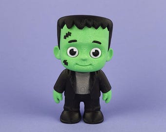 May include: A green Frankenstein's monster figure with black hair, a black suit, and a grey shirt. The figure has stitches on its face and large white eyes. The background is a solid purple colour.