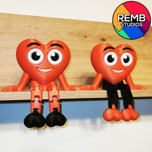 Puede incluir: Dos figuritas rojas en forma de coraz&oacute;n con caras sonrientes y ojos grandes est&aacute;n sentadas en un estante de madera. Cada figurita tiene piernas articuladas negras.