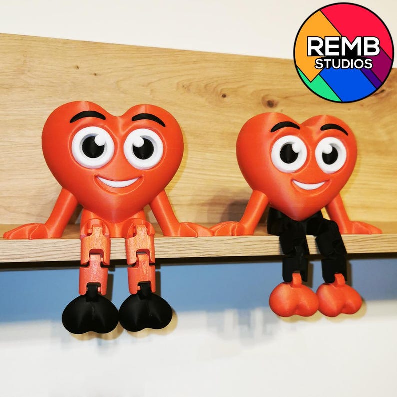 Puede incluir: Dos figuritas rojas en forma de coraz&oacute;n con caras sonrientes y ojos grandes est&aacute;n sentadas en un estante de madera. Cada figurita tiene piernas articuladas negras.