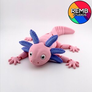 May include: A pink axolotl figurine with blue gills and articulated limbs. The toy has a detailed design with visible eye sockets and a textured body. The axolotl is lying flat, with its limbs spread out. The REMB STUDIOS logo is in the upper right corner.
