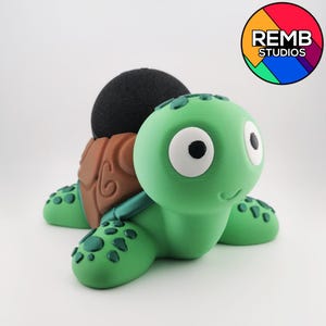 Puede incluir: Un cojín alfiletero caprichoso con forma de tortuga. El cojín tiene un cuerpo verde, un caparazón marrón y una parte superior de tela negra. La tortuga tiene ojos grandes con bordes blancos y una expresión sonriente. El cojín está diseñado para colocarse sobre una superficie plana.