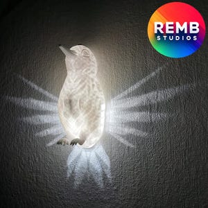 K&ouml;nnte beinhalten: Eine wei&szlig;e Keramik-Pinguin-Nachtlampe mit strukturierter Oberfl&auml;che. Das Licht strahlt ein sanftes Leuchten ab und erzeugt einen Schatten von Fl&uuml;geln an der Wand dahinter.