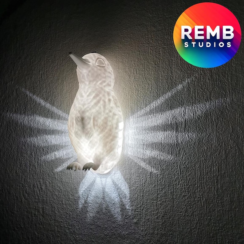 K&ouml;nnte beinhalten: Eine wei&szlig;e Keramik-Pinguin-Nachtlampe mit strukturierter Oberfl&auml;che. Das Licht strahlt ein sanftes Leuchten ab und erzeugt einen Schatten von Fl&uuml;geln an der Wand dahinter.