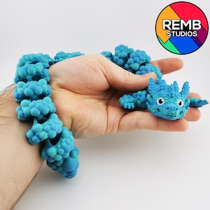 Puede incluir: Un brazalete azul impreso en 3D con un diseño de dragón. El brazalete está hecho de pequeñas cuentas redondas que están conectadas entre sí. El dragón tiene un cuerpo azul con ojos blancos y una lengua rosa. El brazalete está siendo sostenido en una mano.