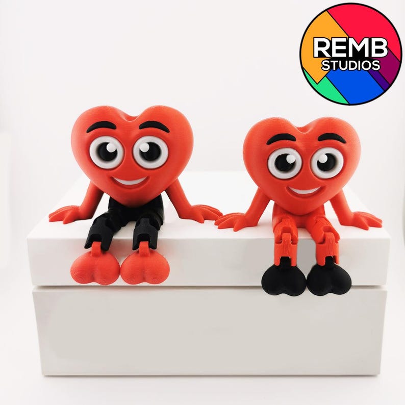 Puede incluir: Dos figuras rojas en forma de coraz&oacute;n con caras sonrientes y ojos grandes est&aacute;n sentadas sobre una superficie blanca. Tienen piernas y pies negros. Las figuras est&aacute;n hechas de un material suave y flexible. El texto "REMB STUDIOS" es visible en un c&iacute;rculo colorido en la esquina superior derecha de la imagen.