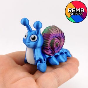 May include: A 3D-printed blue snail figurine with a white underbelly and a multi-colored shell. The snail has large, expressive eyes and is posed on a hand. The logo for REMB STUDIOS is in the upper right corner.
