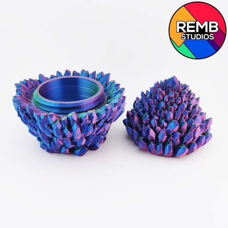 May include: Two-piece iridescent crystal-shaped container. The container is open, revealing a hollow interior. The lid is separate and also crystal-shaped. The colors shift from blue to purple and pink. The container is on a white background.