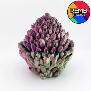 May include: A 3D-printed sculpture of a cluster of crystals in shades of purple, green, and blue. The crystals are arranged in a layered, geometric pattern, creating a textured surface. The sculpture has a rounded base and a pointed top. The REMB STUDIOS logo is in the upper right corner.