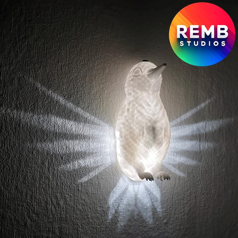 K&ouml;nnte beinhalten: Eine wei&szlig;e Keramik-Pinguin-Nachtlampe mit strukturierter Oberfl&auml;che. Das Licht ist an und wirft einen warmen Schein auf die Wand dahinter.