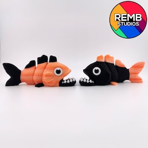 Può includere: Due pesci giocattolo, uno arancione e nero, l'altro nero e arancione, sono esposti fianco a fianco. Ogni pesce ha una bocca spalancata con denti bianchi e un grande occhio rotondo. I pesci sono su una superficie bianca.