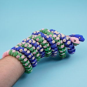 May include: A snake-shaped bracelet in blue, green, and pink. The bracelet features flower-shaped details. The snake's head is blue and pink. The bracelet is worn on a wrist against a light blue background.