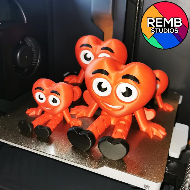 Puede incluir: Tres figuras de coraz&oacute;n rojo impresas en 3D con caras sonrientes y ojos grandes. Las figuras est&aacute;n sentadas sobre una superficie blanca con un fondo negro. La figura m&aacute;s grande tiene el texto "REMB STUDIOS" impreso en ella.