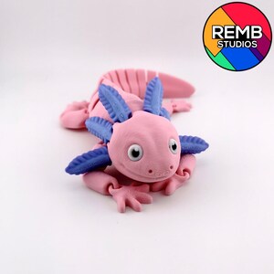 May include: A pink axolotl figurine with blue frills and limbs. The axolotl has large, round eyes and is lying on a white surface. The REMB STUDIOS logo is in the upper right corner.