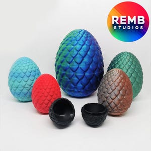 May include: A set of six dragon egg-shaped containers in various colours, including blue, green, red, and brown. The eggs are made of a textured material and have a glossy finish. The eggs are arranged in a semi-circle on a white surface. REMB STUDIOS is printed on a rainbow background.