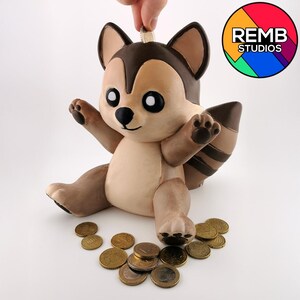 Op de afbeelding: Een bruin en tan wasbeer spaarpot met een gleuf bovenop voor munten. De wasbeer zit op een wit oppervlak met verschillende gouden munten eromheen verspreid.