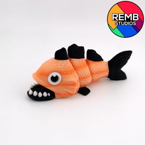 Può includere: Una stravagante statuetta a forma di pesce arancione e nero con la bocca spalancata che rivela i denti bianchi. Il pesce ha un motivo a strisce sul corpo e un occhio grande. La coda e le pinne sono nere. Il logo REMB STUDIOS è visibile.