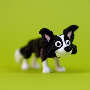 May include: A small, black and white Border Collie figurine with a friendly expression. The dog has large, expressive eyes, perky ears, and white paws. The figurine is set against a bright green background.