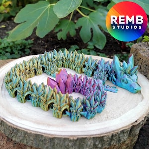 May include: A 3D printed dragon figurine with a crystal-like texture. The dragon is iridescent with shades of blue, green, and purple. The dragon is lying on a wooden surface. REMB STUDIOS is printed on a rainbow circle in the background.