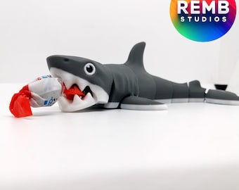 May include: A gray and white shark toy with a candy in its mouth. The candy is wrapped in red and white paper. The toy is on a white surface. REMB STUDIOS is printed on a colorful circle in the background.