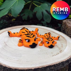 Könnte beinhalten: Zwei orangefarbene und schwarze Tigerfiguren auf einer Holzoberfläche. Der größere Tiger liegt auf dem Bauch mit ausgestreckten Beinen. Der kleinere Tiger liegt auf dem Bauch mit gebeugten Beinen.