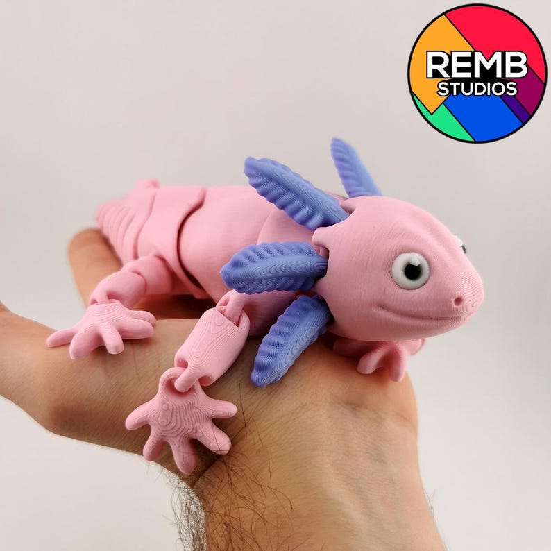 May include: A pink articulated axolotl toy with blue gills and limbs. The toy is held in a hand. The axolotl has a detailed, textured design. The REMB STUDIOS logo is in the upper right corner.