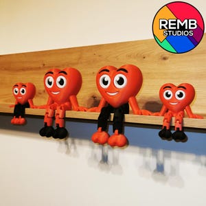Puede incluir: Cuatro figuritas rojas en forma de coraz&oacute;n con caras sonrientes y piernas negras est&aacute;n sentadas en un estante de madera. Las figuritas son de diferentes tama&ntilde;os y est&aacute;n dispuestas de la m&aacute;s grande a la m&aacute;s peque&ntilde;a. La figurita m&aacute;s grande est&aacute; en el centro del estante. El estante est&aacute; contra una pared blanca. El texto "REMB STUDIOS" es visible en un c&iacute;rculo colorido en la esquina superior derecha de la imagen.