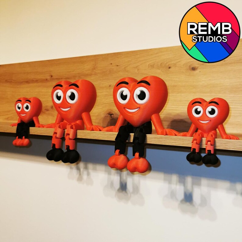 Puede incluir: Cuatro figuritas rojas en forma de coraz&oacute;n con caras sonrientes y piernas negras est&aacute;n sentadas en un estante de madera. Las figuritas son de diferentes tama&ntilde;os y est&aacute;n dispuestas de la m&aacute;s grande a la m&aacute;s peque&ntilde;a. La figurita m&aacute;s grande est&aacute; en el centro del estante. El estante est&aacute; contra una pared blanca. El texto "REMB STUDIOS" es visible en un c&iacute;rculo colorido en la esquina superior derecha de la imagen.