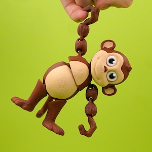 Può includere: Un giocattolo a forma di scimmia articolata in legno con segmenti marroni e beige, sospeso su uno sfondo verde. Una mano tiene il segmento superiore. La faccia della scimmia ha grandi occhi rotondi e piccole orecchie.