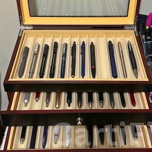 May include: A wooden pen display case with a glass top, showcasing a collection of various pens. The case has multiple compartments lined with beige fabric, holding pens of different colors and designs. The case is open, revealing two levels of pens.