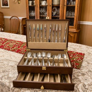 May include: A wooden silverware chest with an open top drawer and a pull-out bottom drawer. The chest is filled with polished silverware, including knives, forks, and spoons. The silverware is arranged neatly in individual slots. The chest is on a table with a patterned tablecloth.
