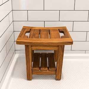 May include: A small, rectangular wooden shower stool with a natural finish. The stool has a slatted top with two hand cutouts and a lower shelf. The stool is made of solid wood and is designed for use in a shower or bathroom.