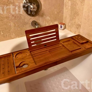 May include: A wooden bathtub tray with multiple compartments and a book rest. The tray is made of a warm-toned wood and features cutouts for a wine glass and other items. It rests on the edge of a white bathtub.