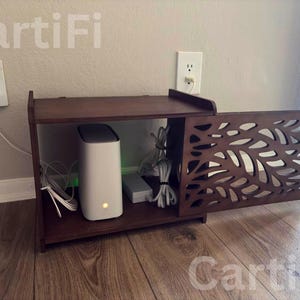 May include: A dark brown wooden cabinet with a decorative leaf-patterned door, housing a white modem and various cables. The cabinet sits on a wooden floor, with a wall outlet and a wall-mounted device visible.