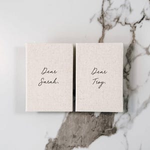 May include: Two rectangular, off-white canvas notebooks with the words "Dear Sarah" and "Dear Troy" in elegant black script. The notebooks rest on a marble surface with gray veining.