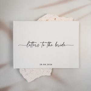May include: A white rectangular book with the text "letters to the bride" in black cursive script centered on the front cover. Below the text, the date "28.08.2026" is printed in a simple black sans-serif font on the lower portion.