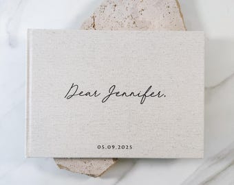 May include: A rectangular, light beige fabric-covered book with a hard cover. The front cover features the black printed text "Dear Jennifer." in a cursive script font, followed by the date "05.09.2025" in a simple, clean sans-serif font.