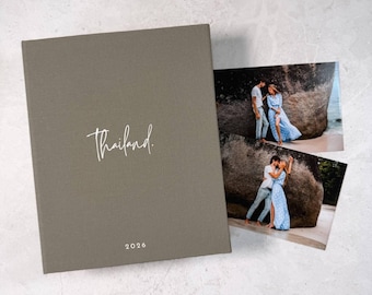 May include: A rectangular photo album with a solid olive-gray fabric cover. The word "Thailand." is printed in white script in the center, with "2026" printed below it. Two rectangular photographs showing a couple on a beach are placed beside it.