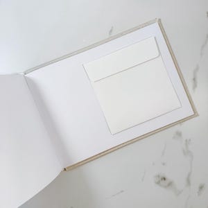 May include: An open, off-white scrapbook with a white envelope attached to the right page. The book's pages are blank, and the cover has a light beige fabric texture. The background is a white marble surface.